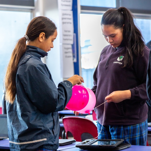 Two young female students at a science fair tying two pink balloons together